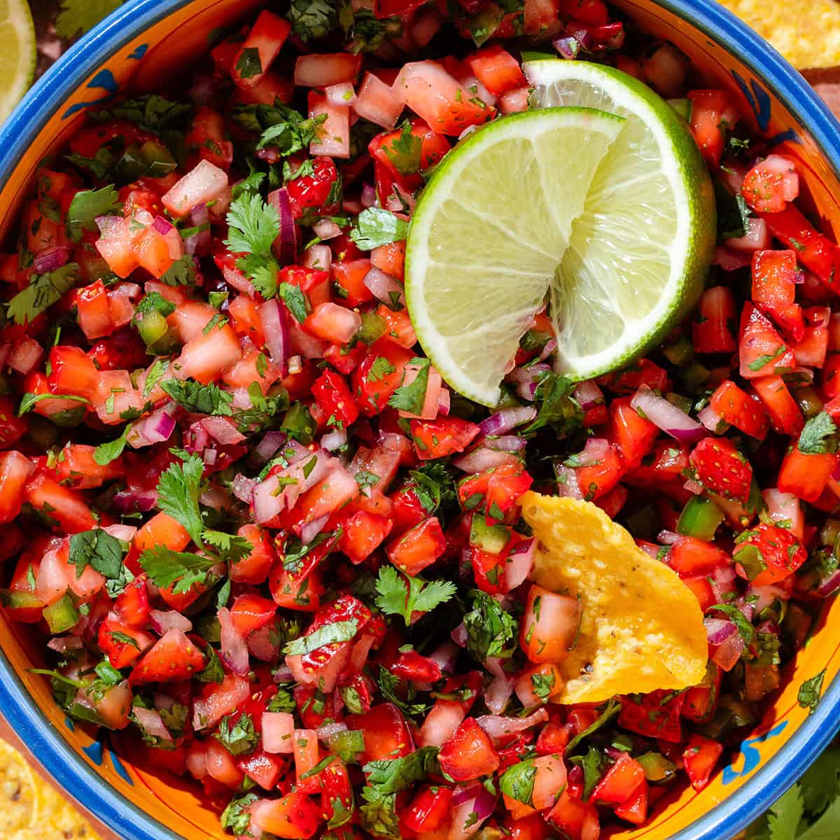 Fresh strawberry salsa in a colorful bowl garnished with two lime wedges and with a tortilla chip dipped into the salsa.