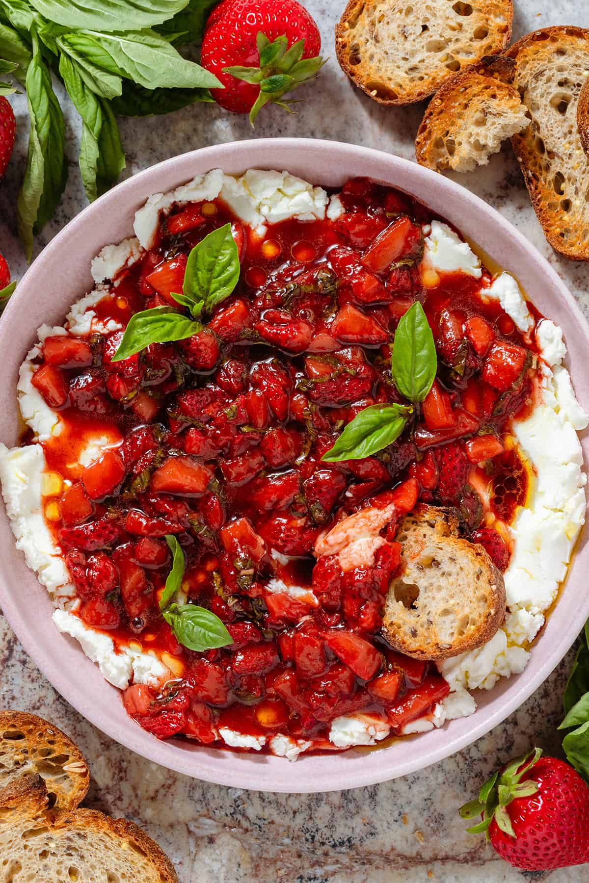 A bowl of soft goat cheese topped with sauteed balsamic strawberries and fresh basil with a slice of toasted baguette dipped into it.