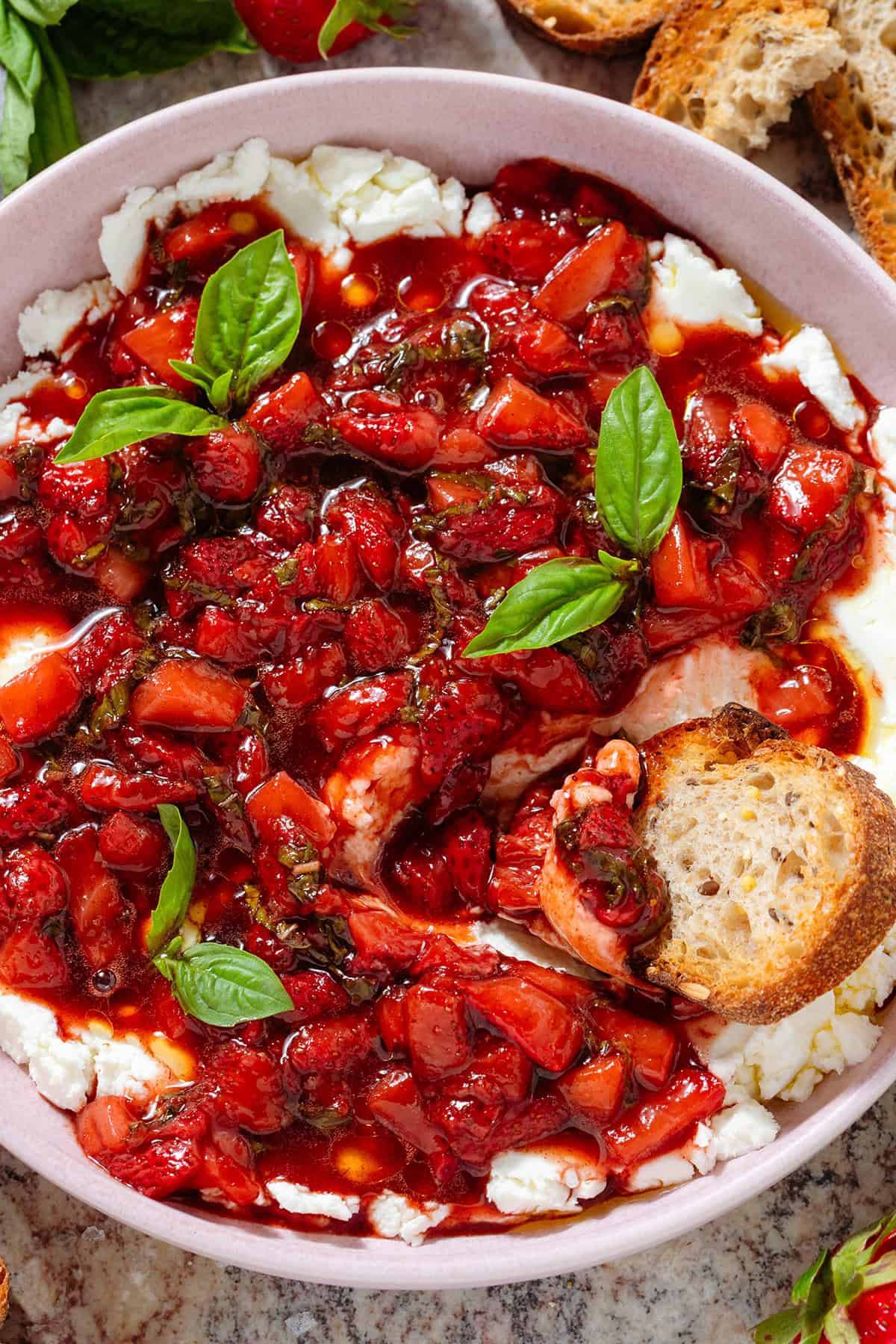 A bowl of soft goat cheese topped with sauteed balsamic strawberries and fresh basil with a slice of toasted baguette dipped into it.