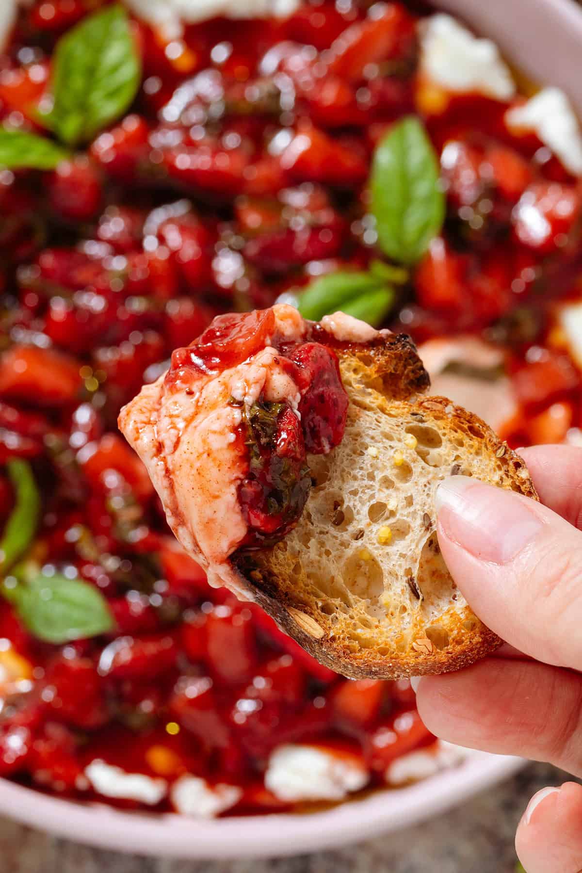 A hand scooping up goat cheese with balsamic strawberries using a slice of toasted baguette.