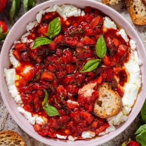 A bowl of soft goat cheese topped with sauteed balsamic strawberries and fresh basil with a slice of toasted baguette dipped into it.