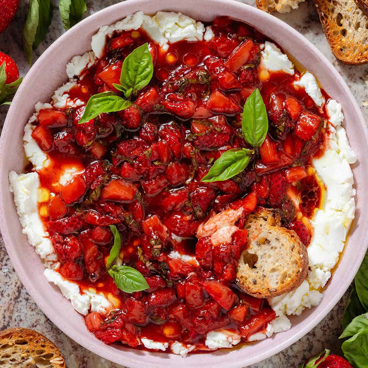 A bowl of soft goat cheese topped with sauteed balsamic strawberries and fresh basil with a slice of toasted baguette dipped into it.