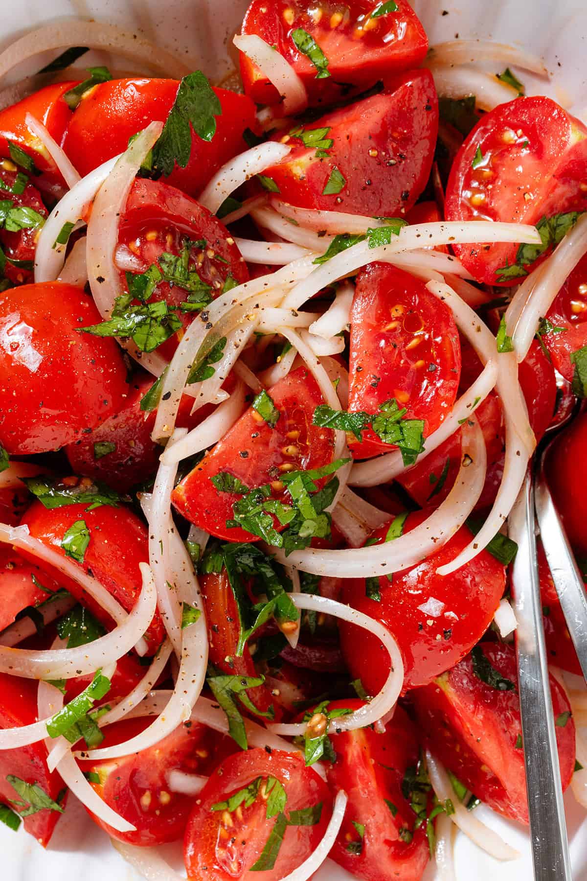 Tomato and onion salad with parsley in a white bowl seasoned with black pepper, with a fork and a spoon in the bowl.