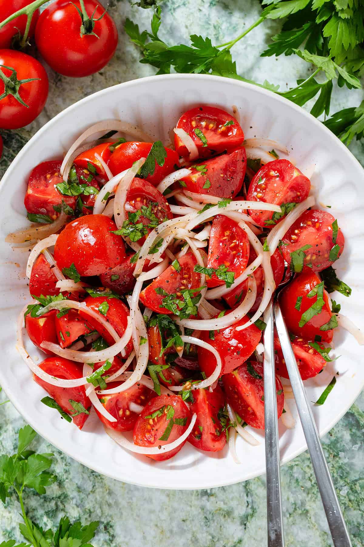 Tomato and onion salad with parsley in a white bowl seasoned with black pepper, with a fork and a spoon in the bowl.