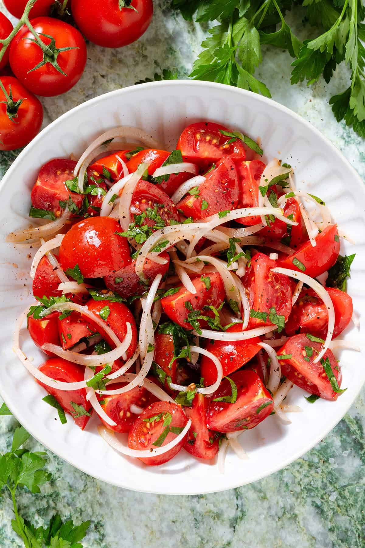 Tomato and onion salad with parsley in a white bowl seasoned with black pepper, with more tomatoes and parsley around the bowl.