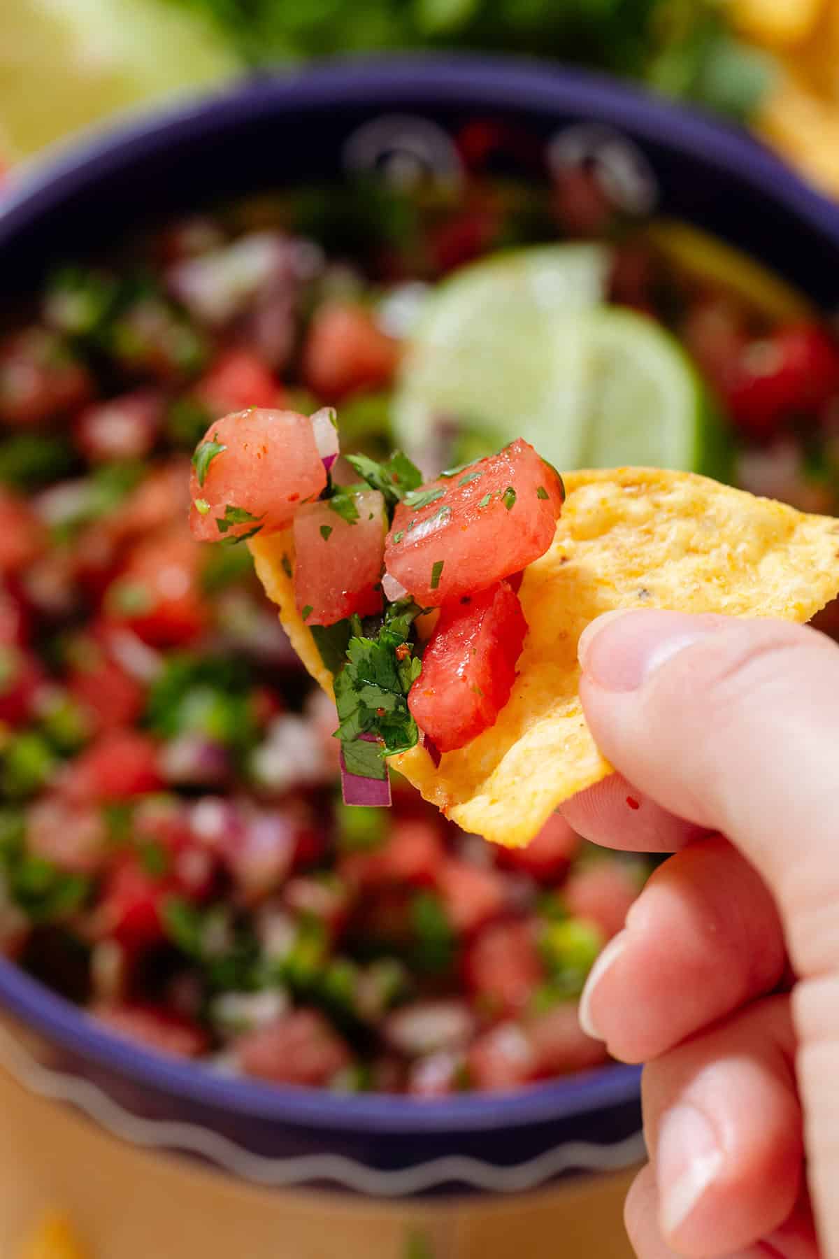 A hand holding a tortilla chip scooping up watermelon salsa over a bowl with more of it.