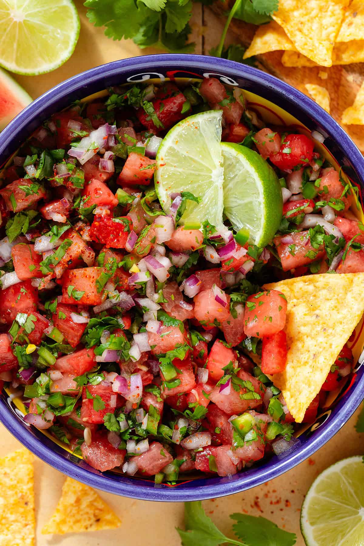 Fresh watermelon salsa in a colorful bowl with a blue rim garnished with two lemon wedges and with a tortilla chip in the bowl.