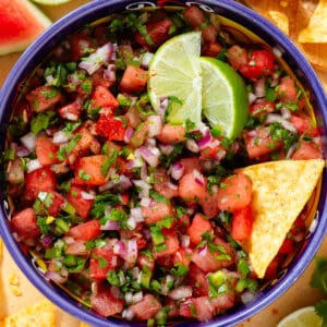 Fresh watermelon salsa in a colorful bowl with a blue rim garnished with two lemon wedges and with a tortilla chip in the bowl.