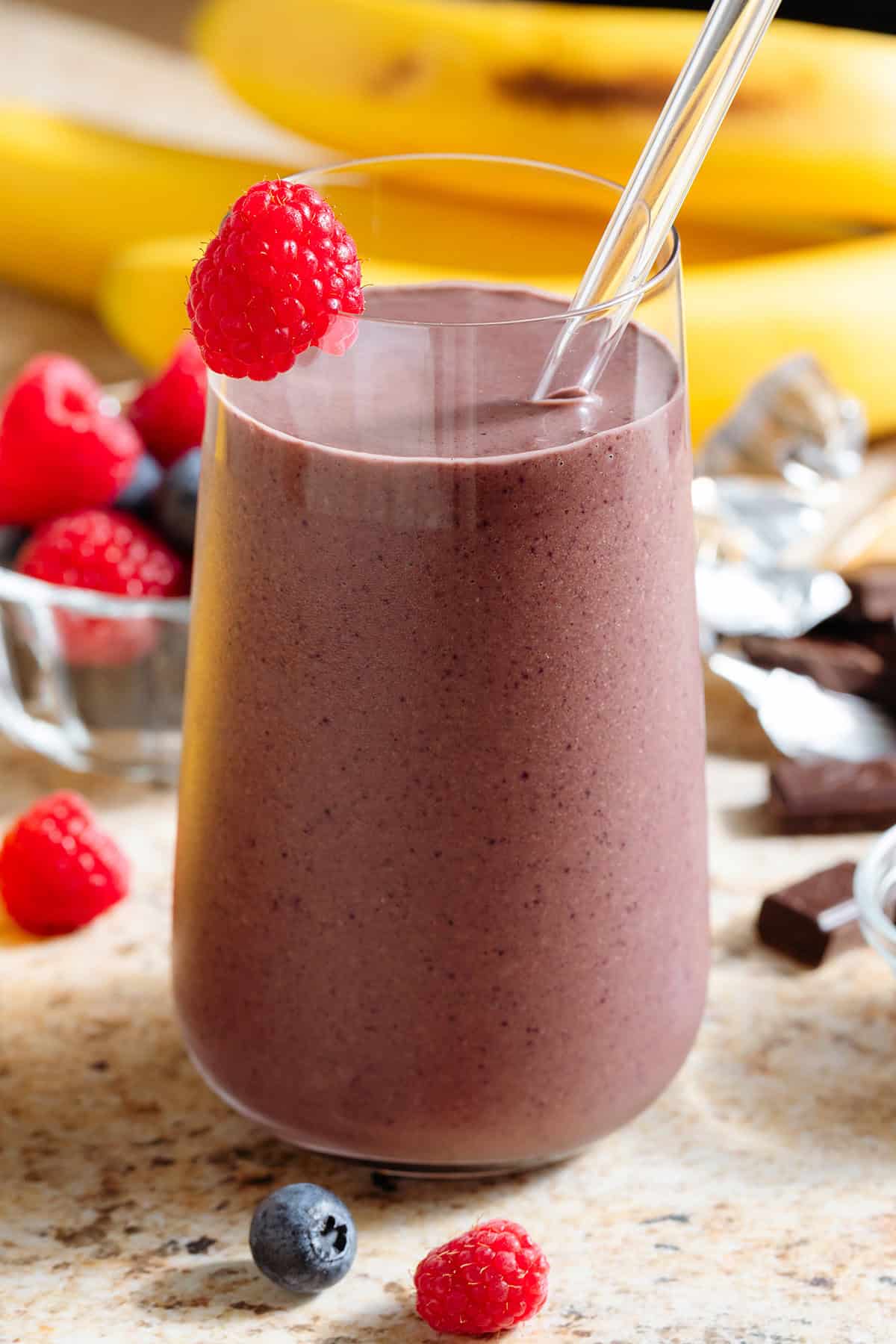 A purple chocolate acai smoothie in a tall glass with a glass straw garnished with a fresh raspberry on the rim with more berries in the background.