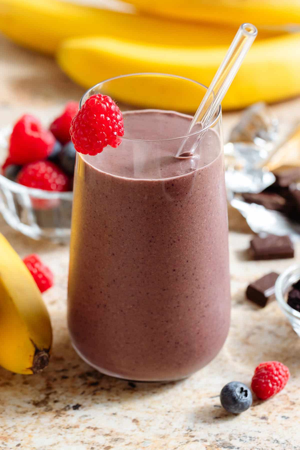 A purple chocolate acai smoothie in a tall glass with a glass straw garnished with a fresh raspberry on the rim.