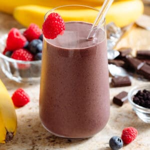 A purple chocolate acai smoothie in a tall glass with a glass straw garnished with a fresh raspberry on the rim.