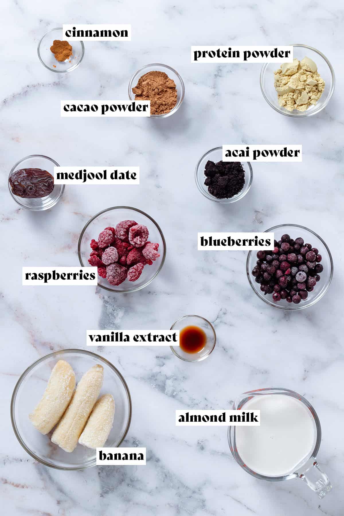 Ingredinets laid out in small glass bowls like frozen bananas, raspberries, bluberries, protein powder, cacao powder, acai powder, vanilla, medjool date, and cinnamon with text overlay.