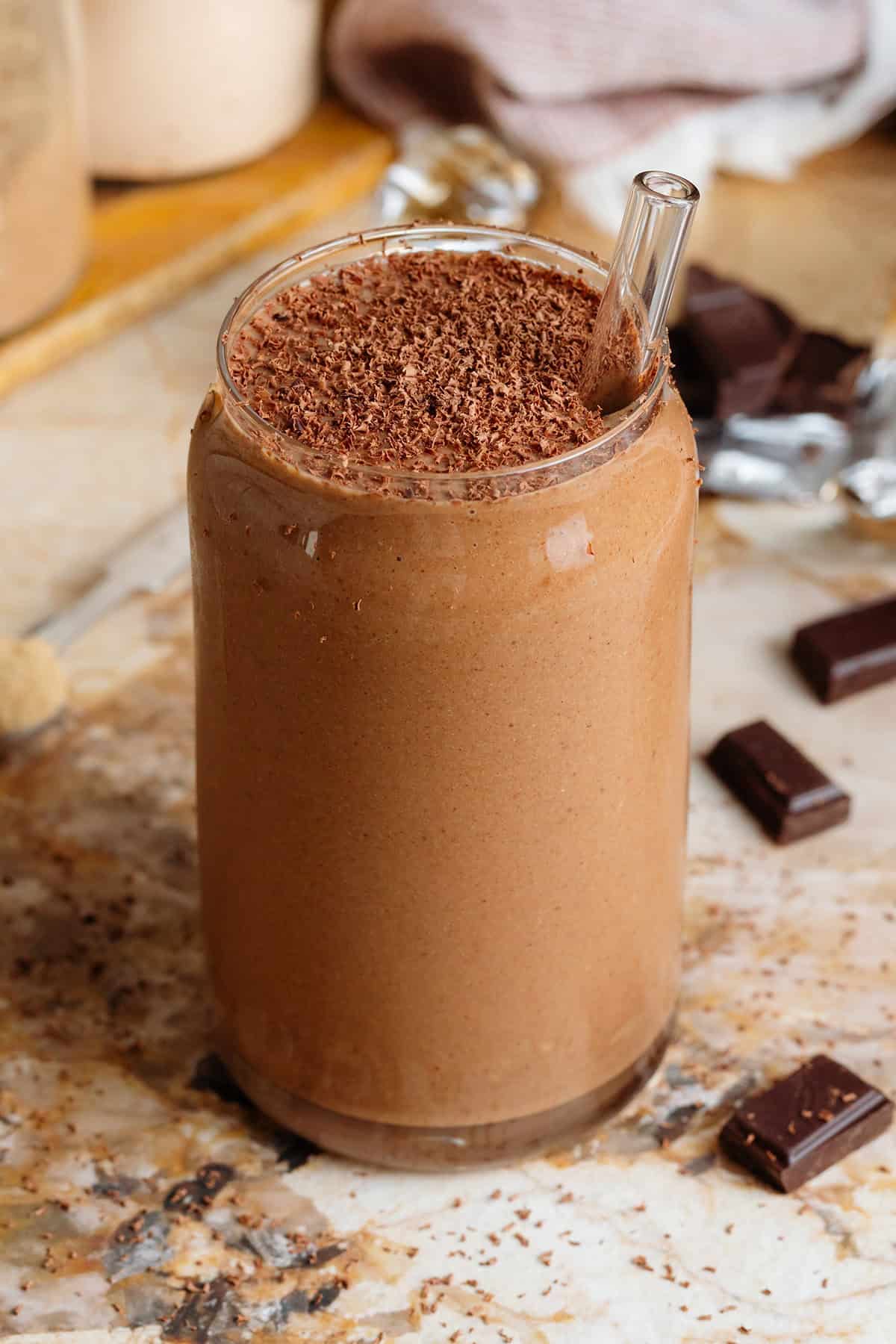 Creamy chocolate smoothie in a glass shaped like a soda can with a glass straw and garnished with grated chocolate with chocolate squares around the glass.