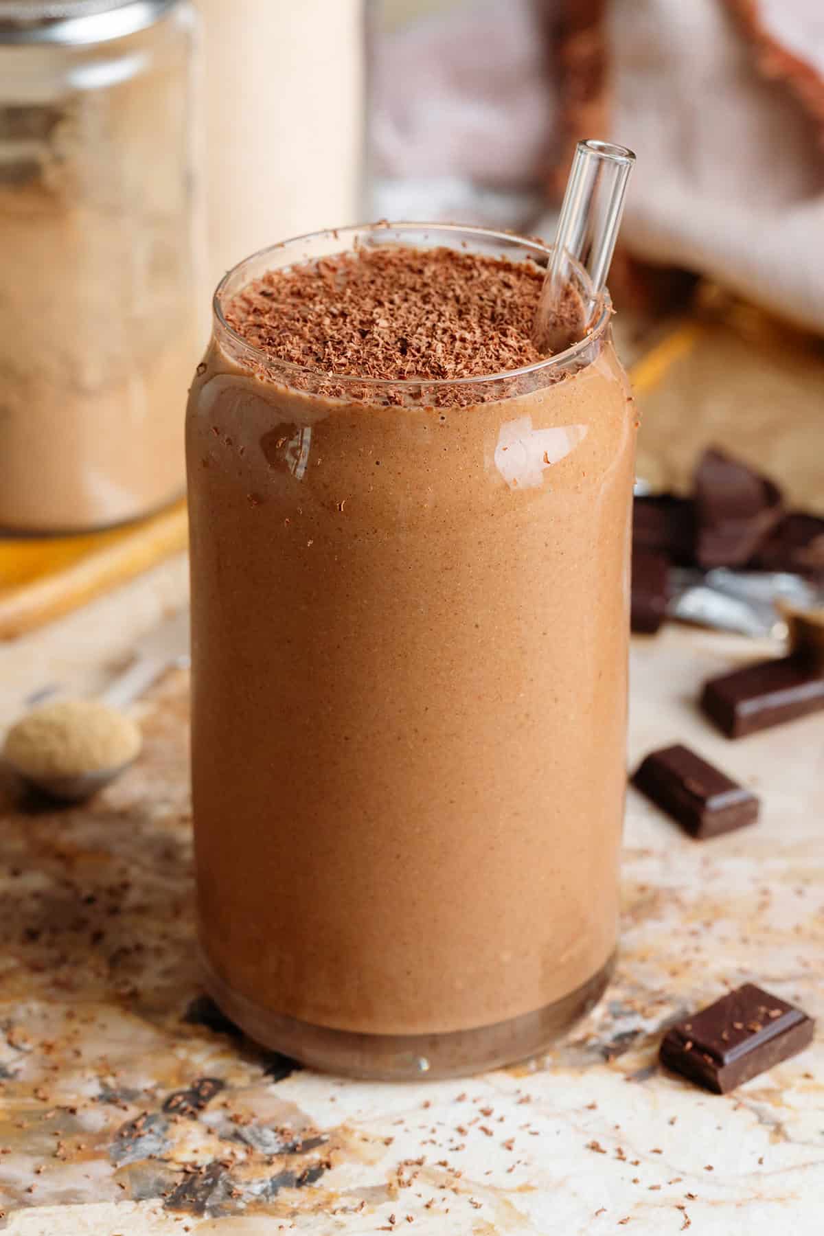 Creamy chocolate smoothie in a glass shaped like a soda can with a glass straw and garnished with grated chocolate with chocolate squares around the glass.