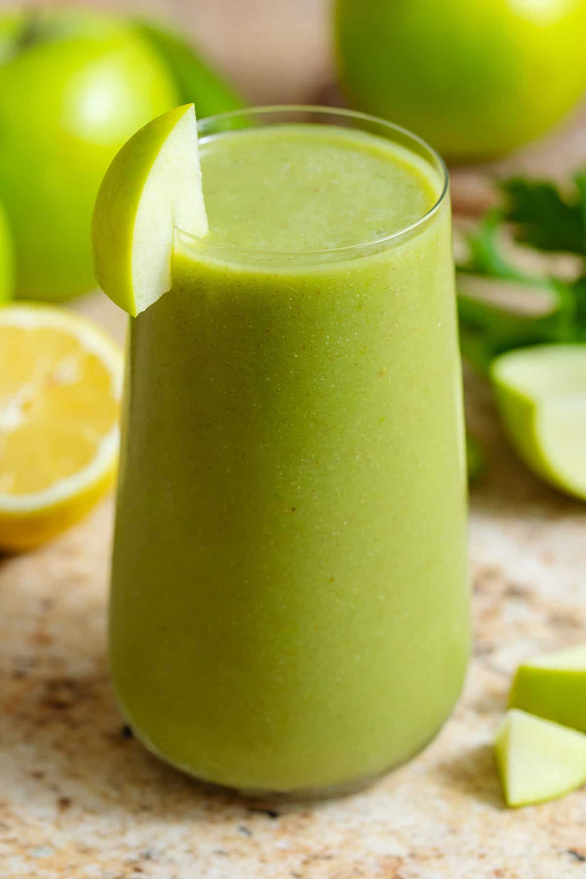 Bright green smoothie in a tall glass garnished with a slice of a green apple with more apples in the background.