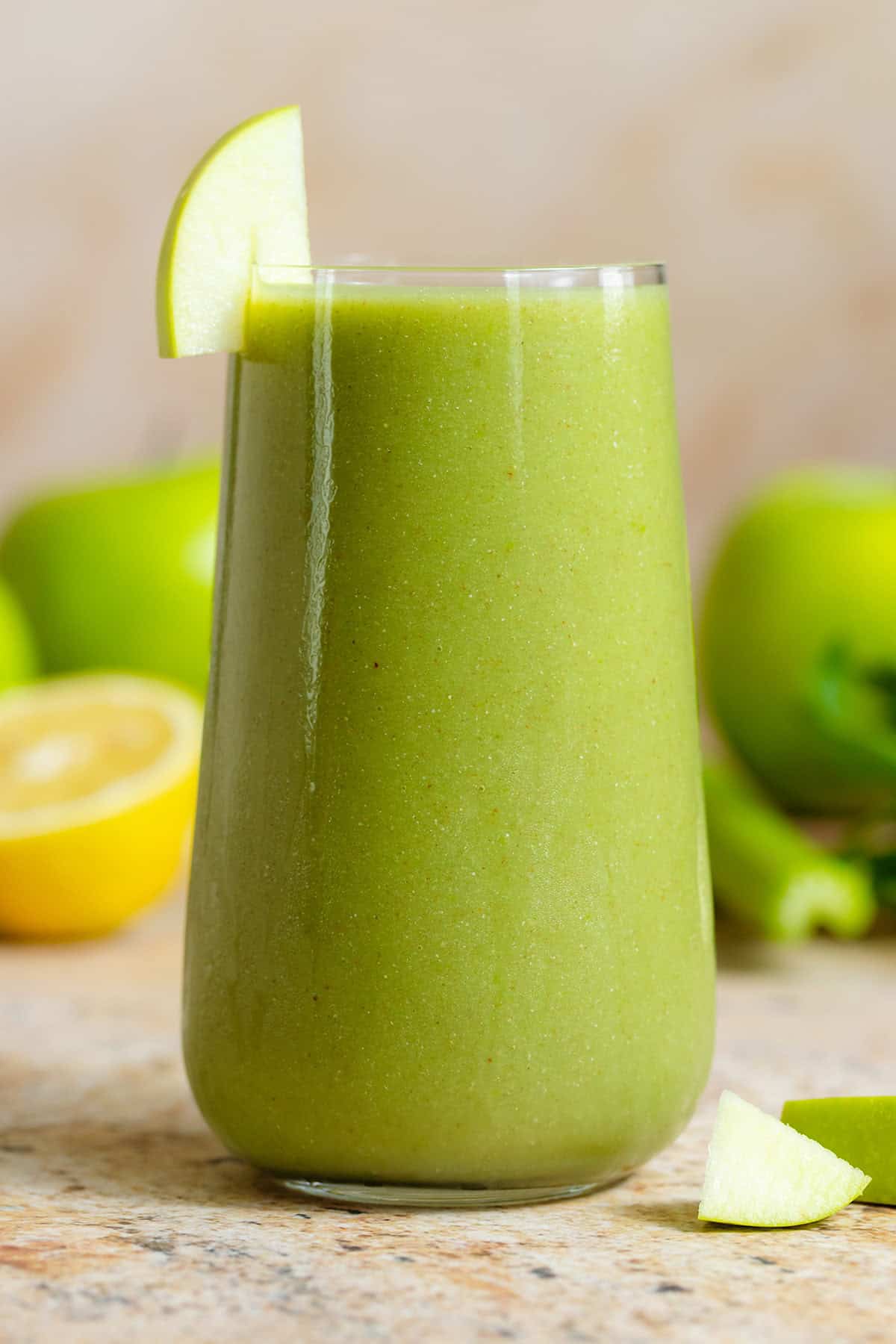 Bright green smoothie in a tall glass garnished with a slice of a green apple with more apples, lemon, and celery in the background.