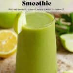 Bright green smoothie in a tall glass garnished with a slice of a green apple with more apples, lemon, and celery in the background.