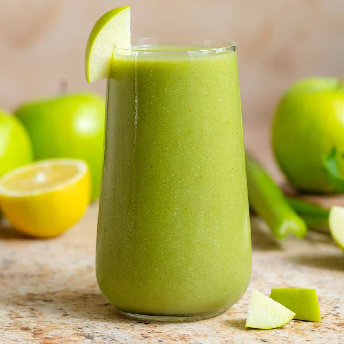 Bright green smoothie in a tall glass garnished with a slice of a green apple with more apples, lemon, and celery in the background.