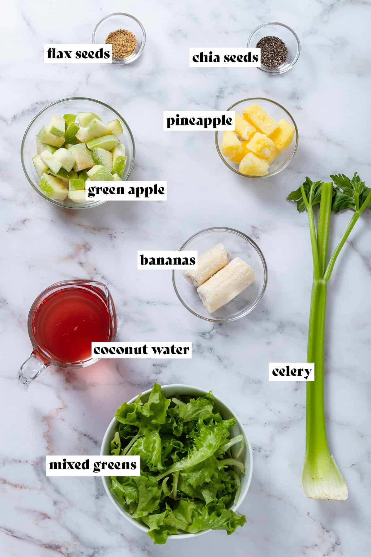 Frozen diced green apple, pineapple, bananas, chia and flax seeds in glass bowls, coconut water in a measuring cup, and a stalk of celery laid out with text overlay.