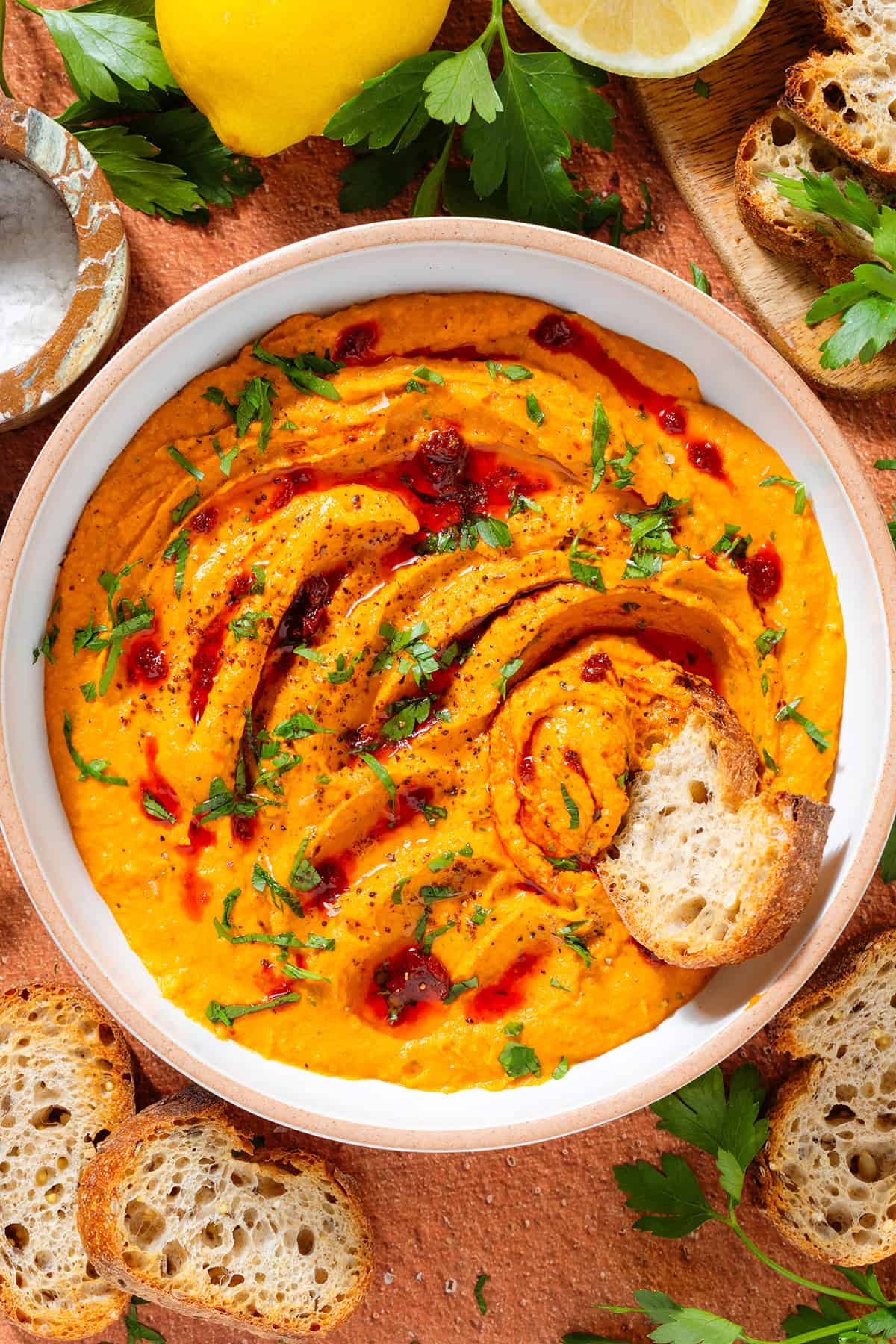 A white bowl with bright orange butter bean harissa dip garnished with chopped parsley, harissa paste, and sumac with a slice of toasted baguette scooping up the dip with more baguette slices around the bowl.
