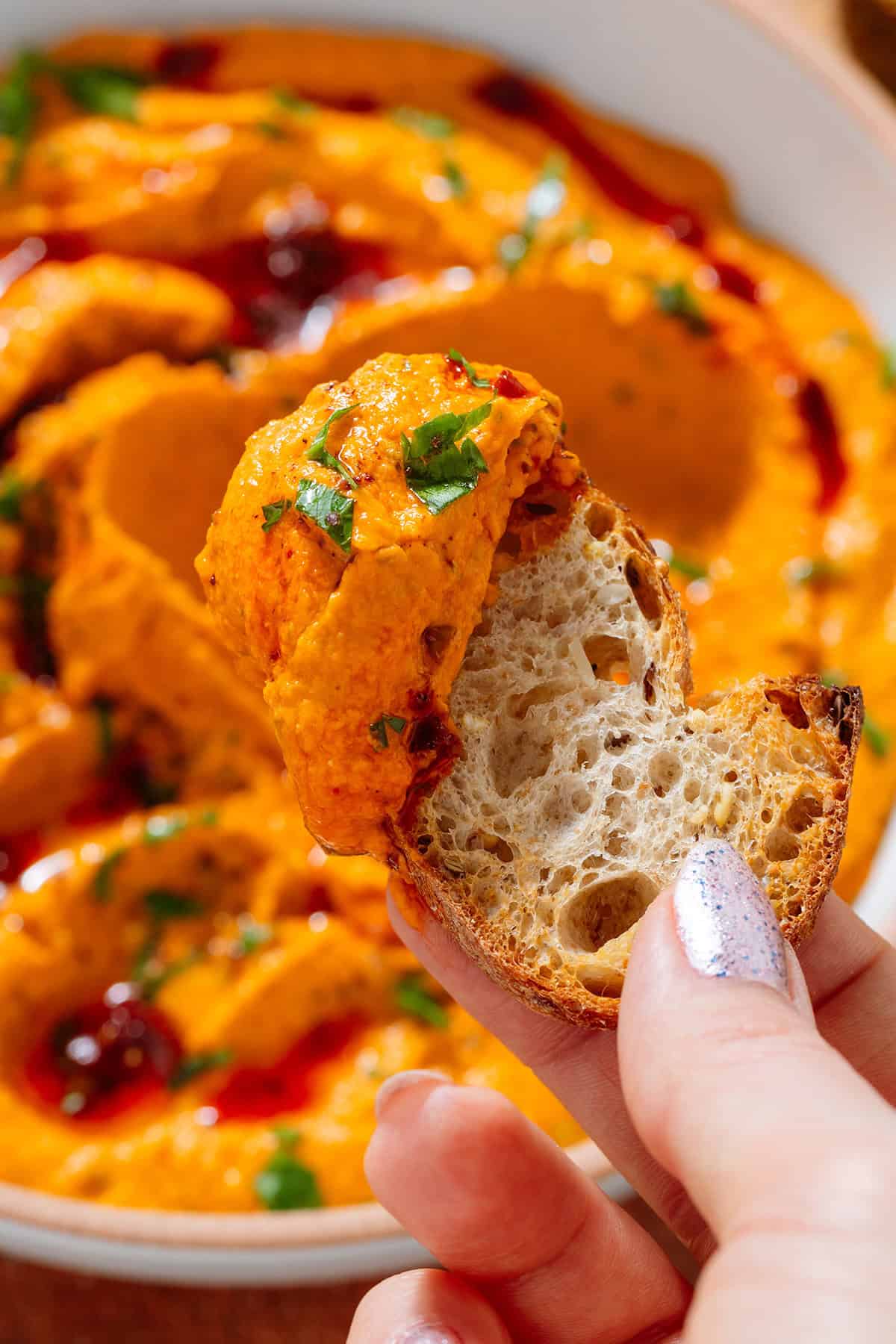A hand holding a slice of baguette with scooped up harissa butter bean dip that's visible behind it.