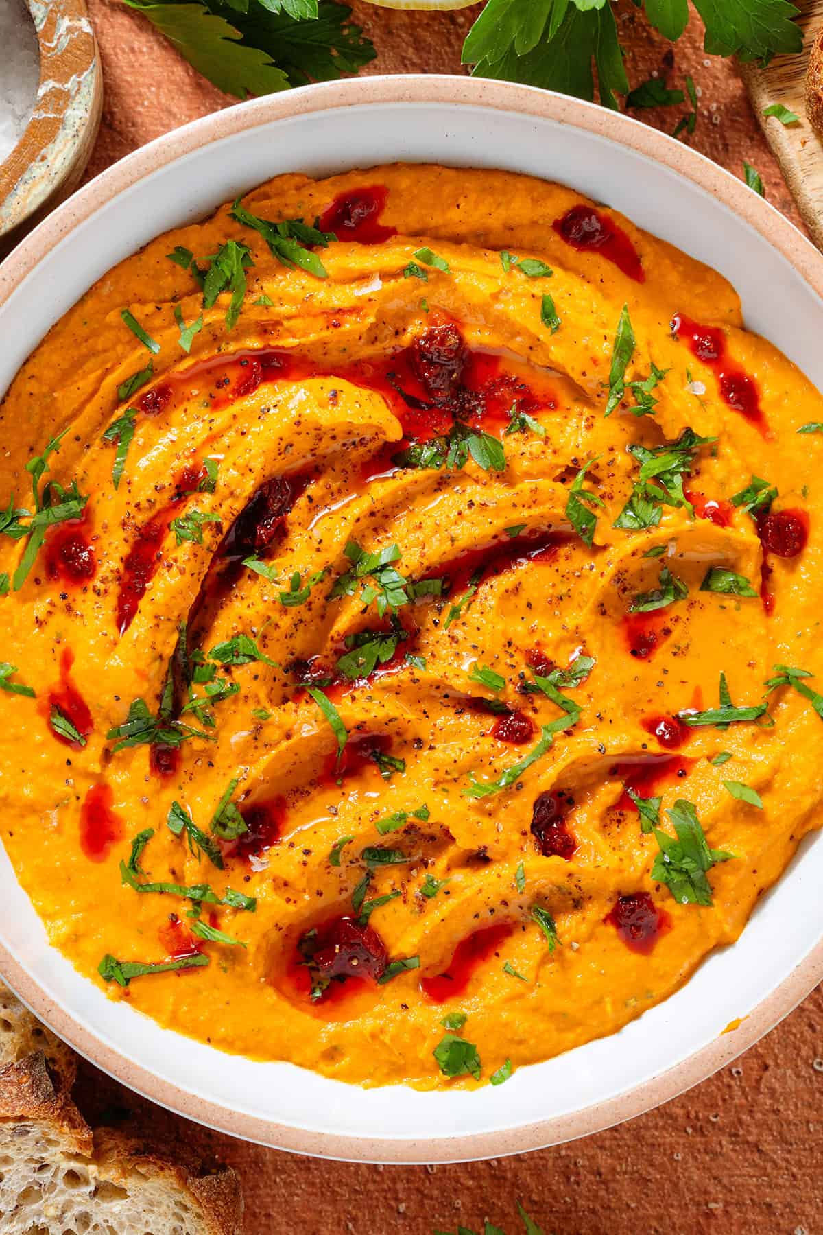 Bright orange butter bean harissa dip garnished with chopped parsley, harissa paste, and sumac in a white bowl with a beige rim.