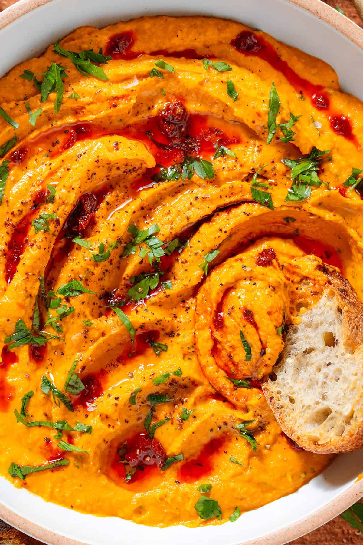 A white bowl with bright orange butter bean harissa dip garnished with chopped parsley, harissa paste, and sumac with a slice of toasted baguette scooping up the dip.