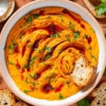 Bright orange butter bean harissa dip garnished with chopped parsley, harissa paste, and sumac in a white bowl with a slice of baguette scooping up the dip.