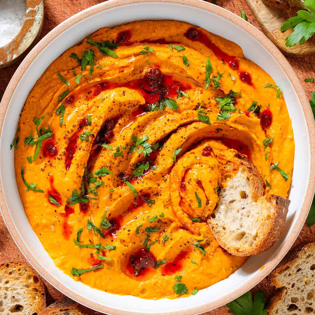 Bright orange butter bean harissa dip garnished with chopped parsley, harissa paste, and sumac in a white bowl with a slice of baguette scooping up the dip.