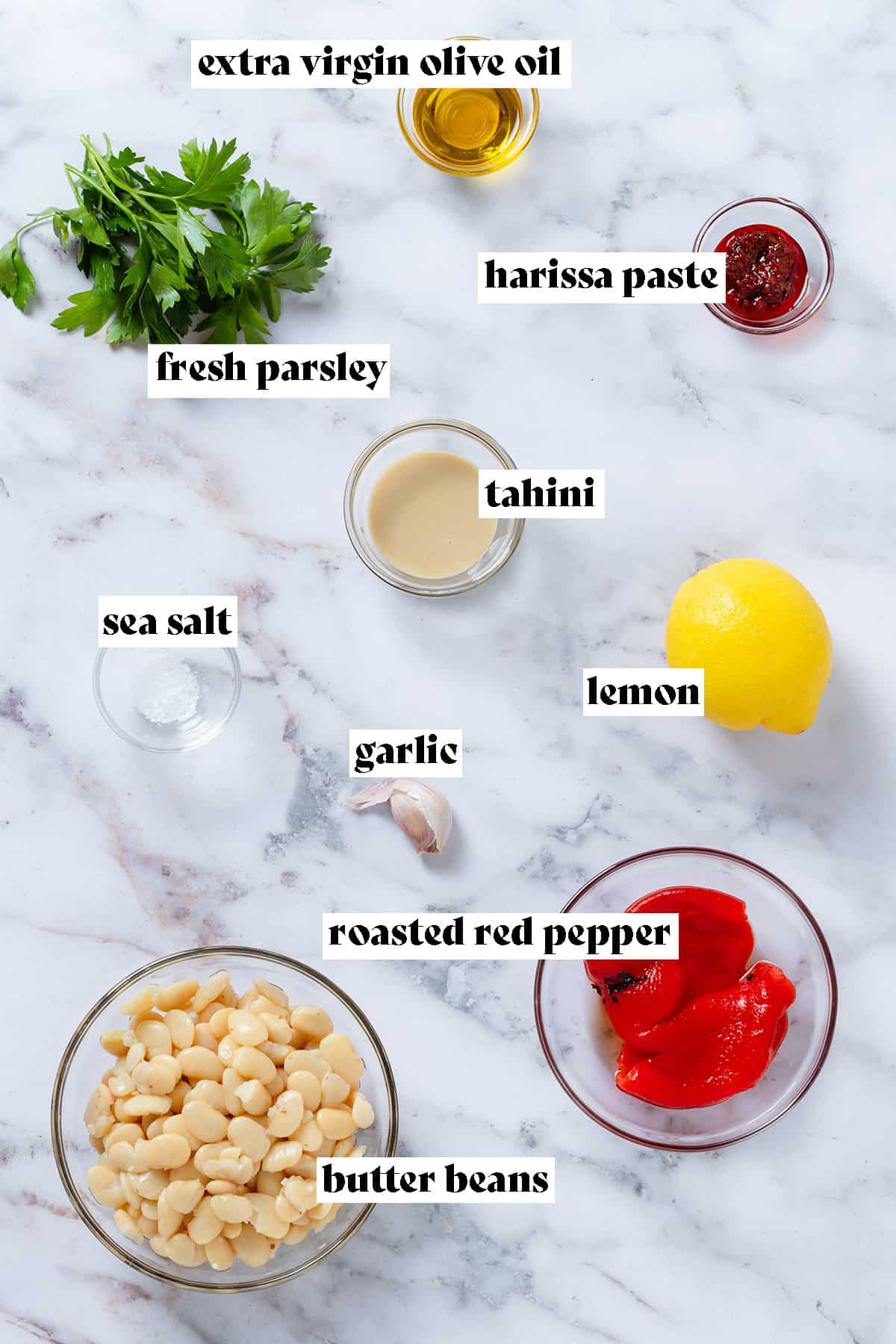Laid out ingredients like butter beans, roasted red pepper, harissa paste, lemon, parsley, and garlic on a grey marble background with text overlay.