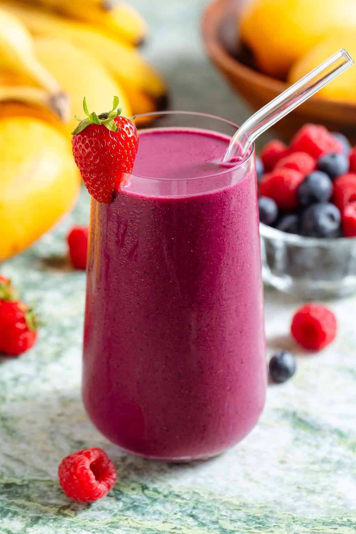 A tall glass with bright purple maqui berry smoothie and a glass straw garnished with a strawberry.