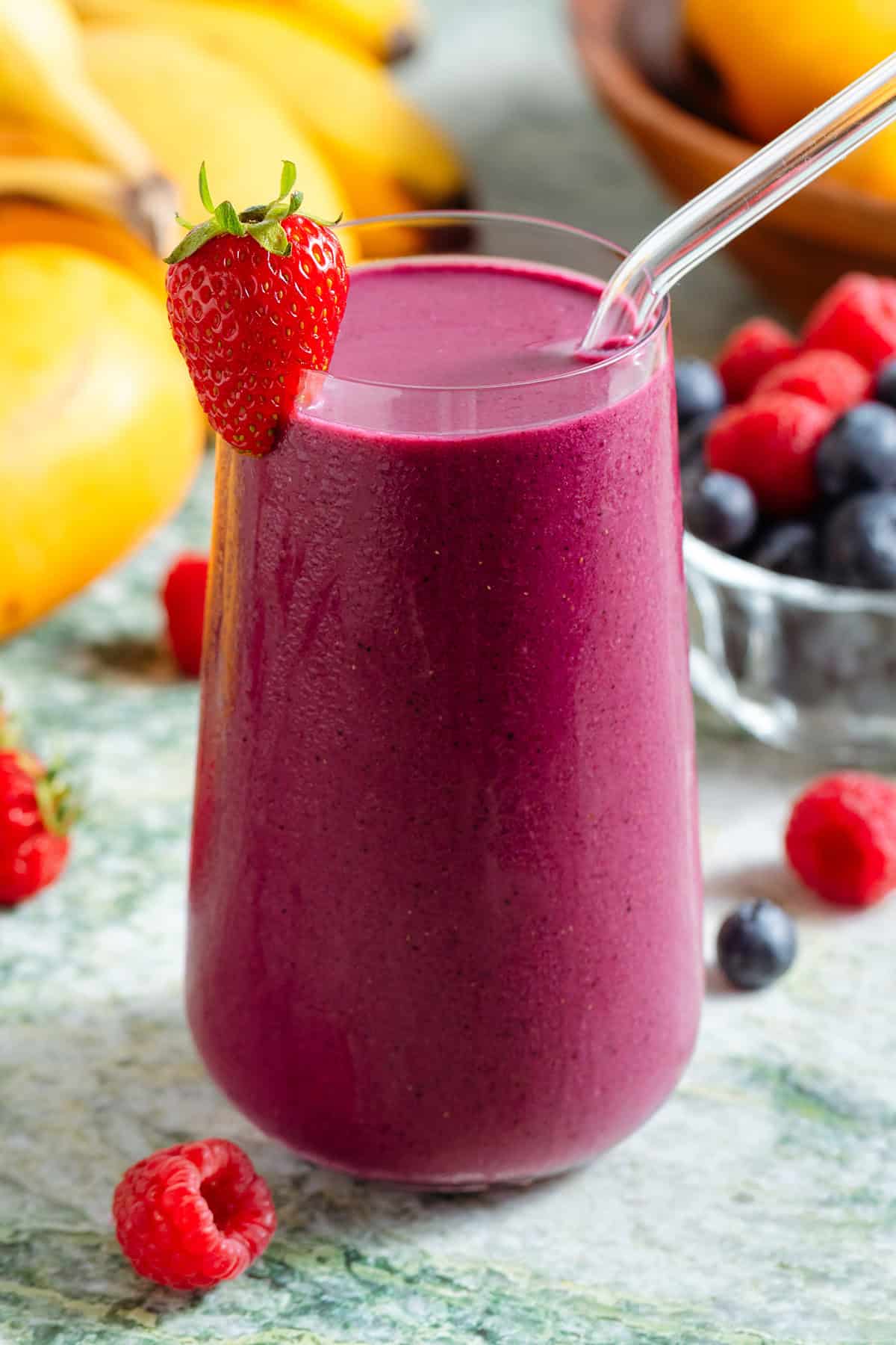 A tall glass with bright purple smoothie and a glass straw garnished with a strawberry and with more berries around the glass.