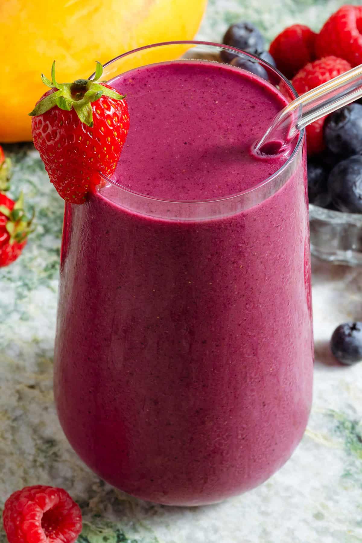 A tall glass with bright purple smoothie and a glass straw garnished with a strawberry and with more berries around the glass.