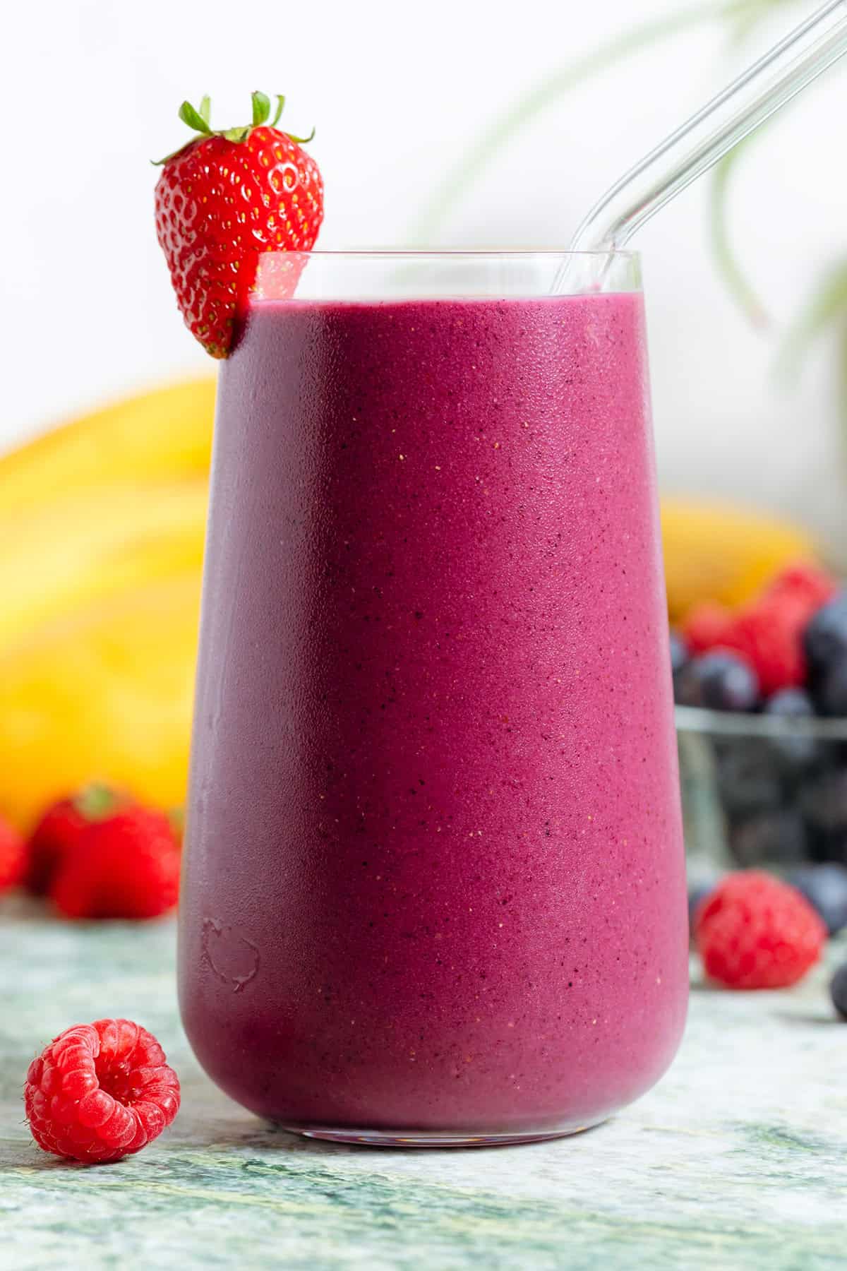 A tall glass with bright purple smoothie and a glass straw garnished with a strawberry and with more berries and bananas around the glass.