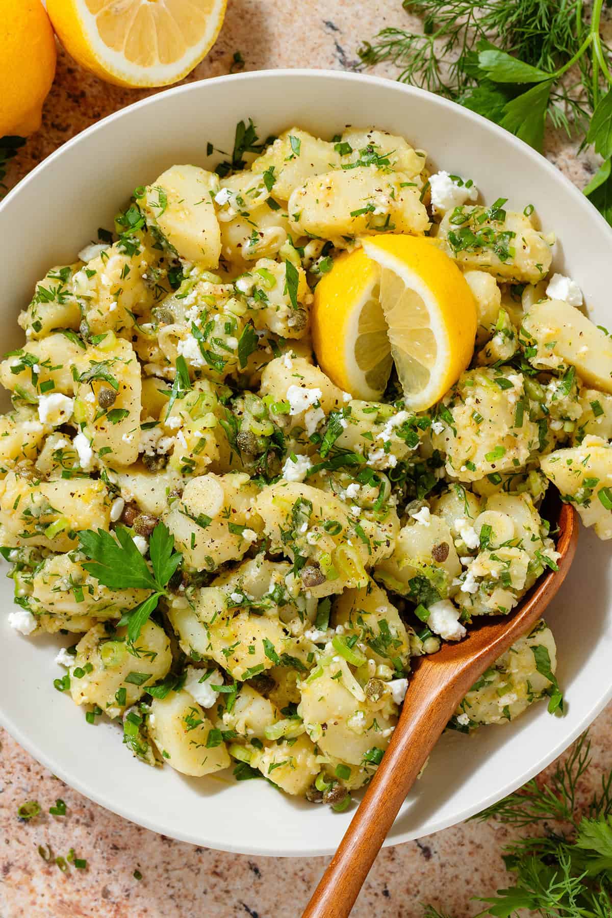 Potato salad with lemon vinaigrette, lots of fresh herbs, and feta in a beige serving bowl garnished with two lemon wedges and with a wooden spoon in the bowl.