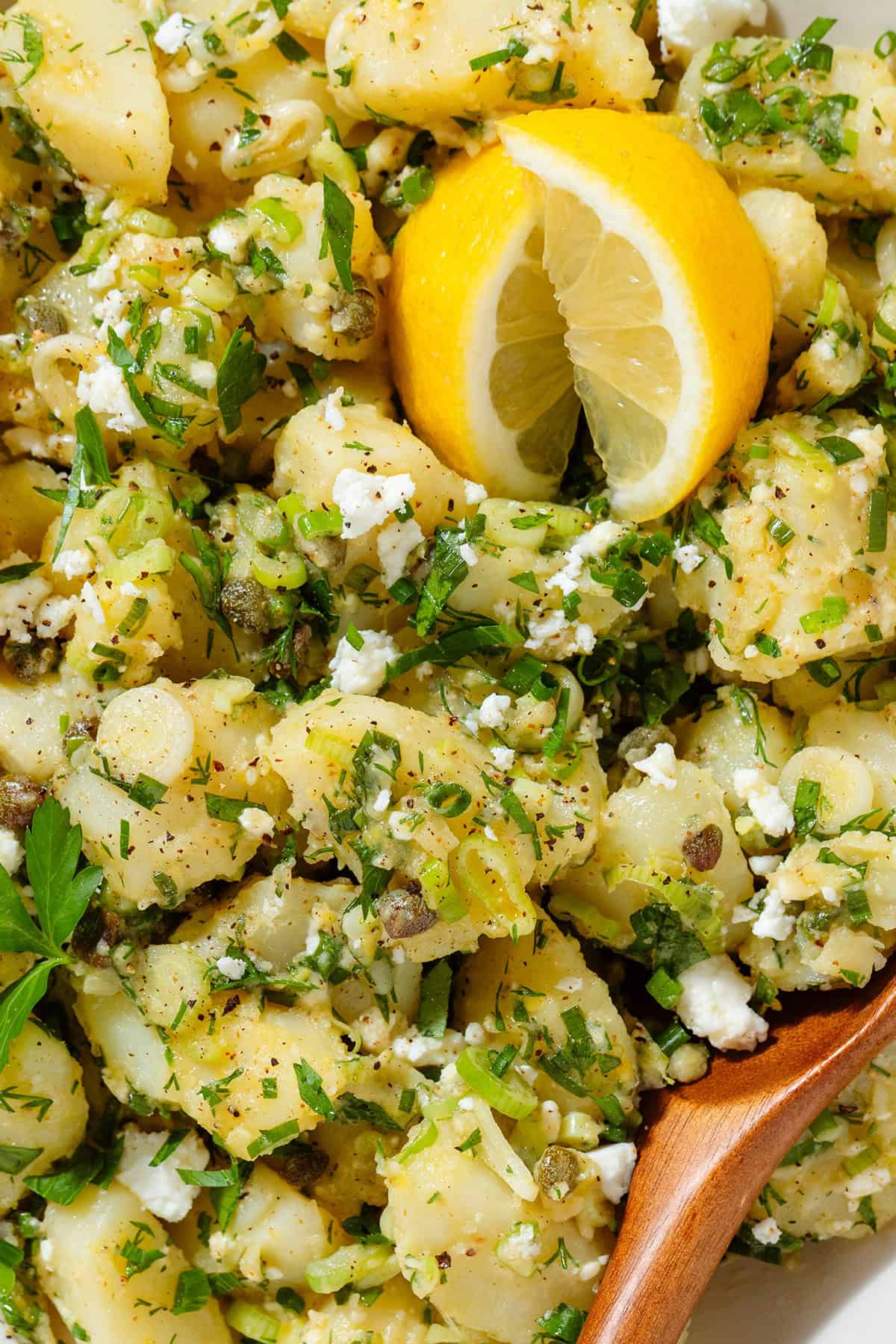 Potato salad with lemon vinaigrette, lots of fresh herbs, and feta in a beige serving bowl garnished with two lemon wedges and with a wooden spoon in the bowl.