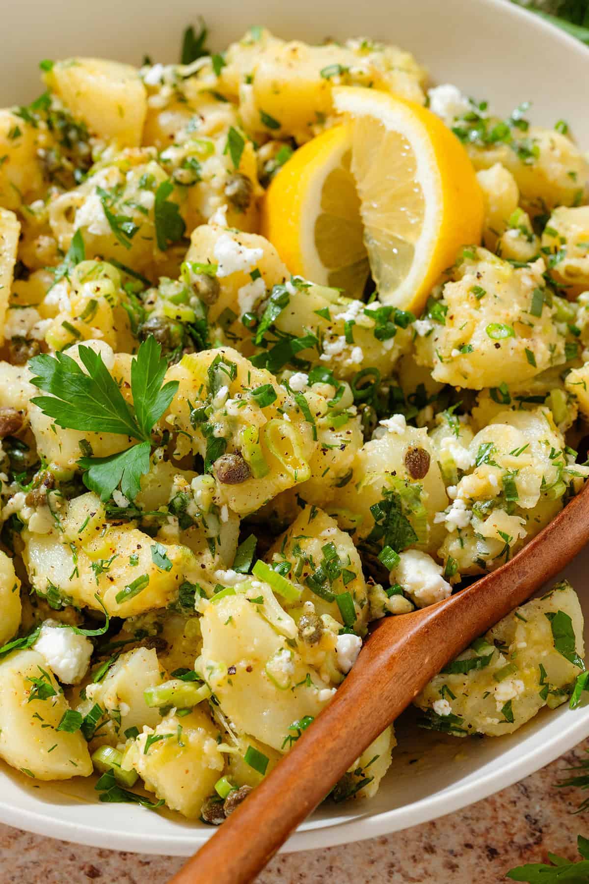 Potato salad with lemon vinaigrette, lots of fresh herbs, and feta in a beige serving bowl garnished with two lemon wedges and with a wooden spoon in the bowl.