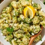 Potato salad with lemon vinaigrette, lots of fresh herbs, and feta in a beige serving bowl garnished with two lemon wedges and more fresh parsley.