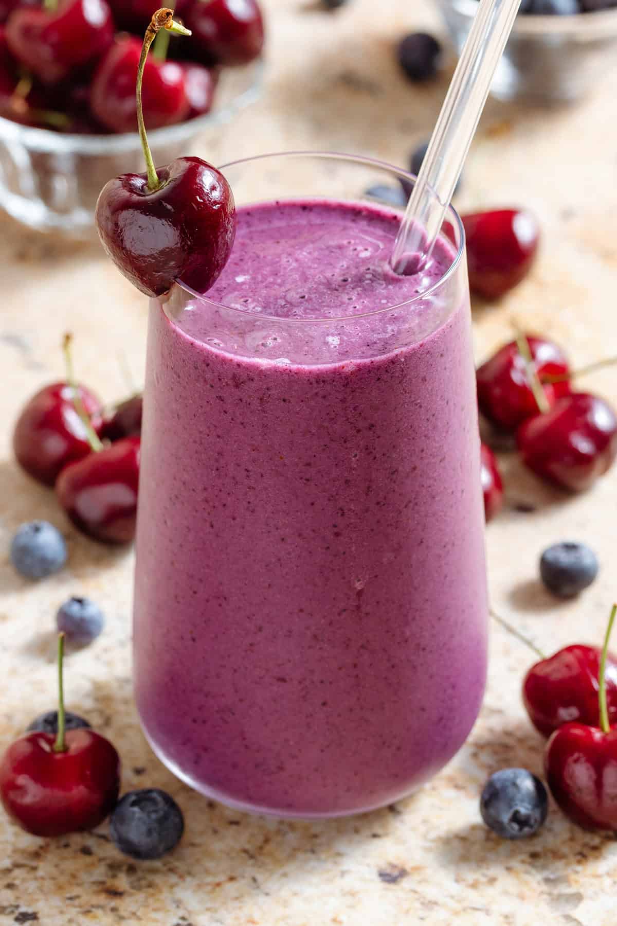 A tall glass with vibrant purple smoothie garnished with a glass straw and cherry on the rim and more cherries and blueberries around the glass.