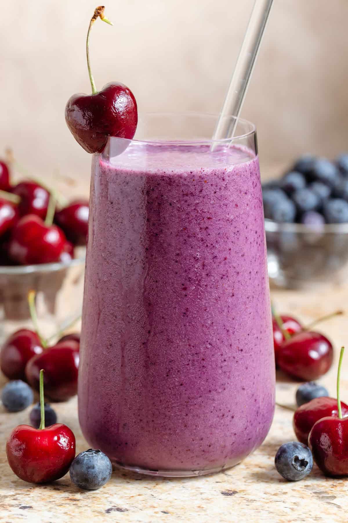A tall glass with vibrant purple smoothie garnished with a glass straw and acherry on the rim and more cherries and blueberries around the glass.