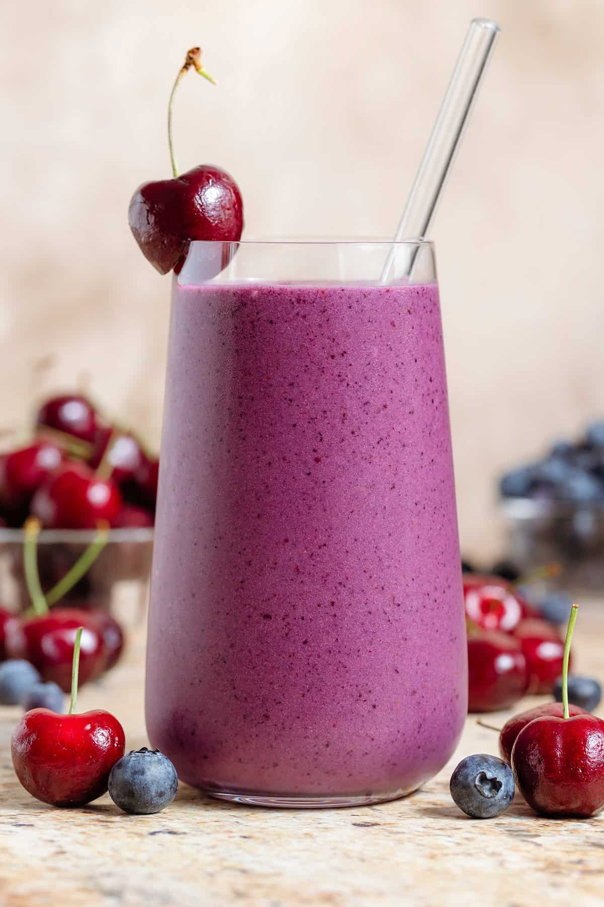 A tall glass with vibrant purple smoothie garnished with a glass straw and cherry on the rim and more cherries and blueberries around the glass.