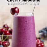 A tall glass with vibrant purple smoothie garnished with a cherry on the rim and a glass straw, more cherries and blueberries around the glass.