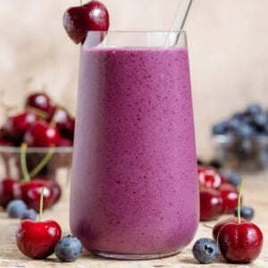 A tall glass with vibrant purple smoothie garnished with a cherry on the rim and a glass straw, more cherries and blueberries around the glass.