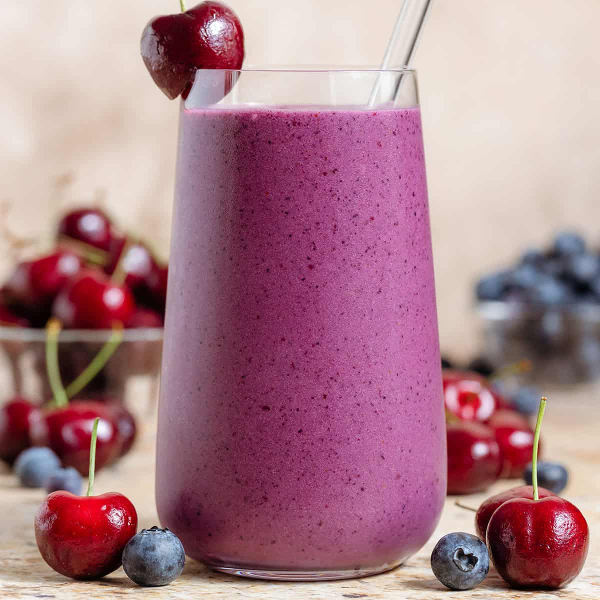 A tall glass with vibrant purple smoothie garnished with a cherry on the rim and a glass straw, more cherries and blueberries around the glass.