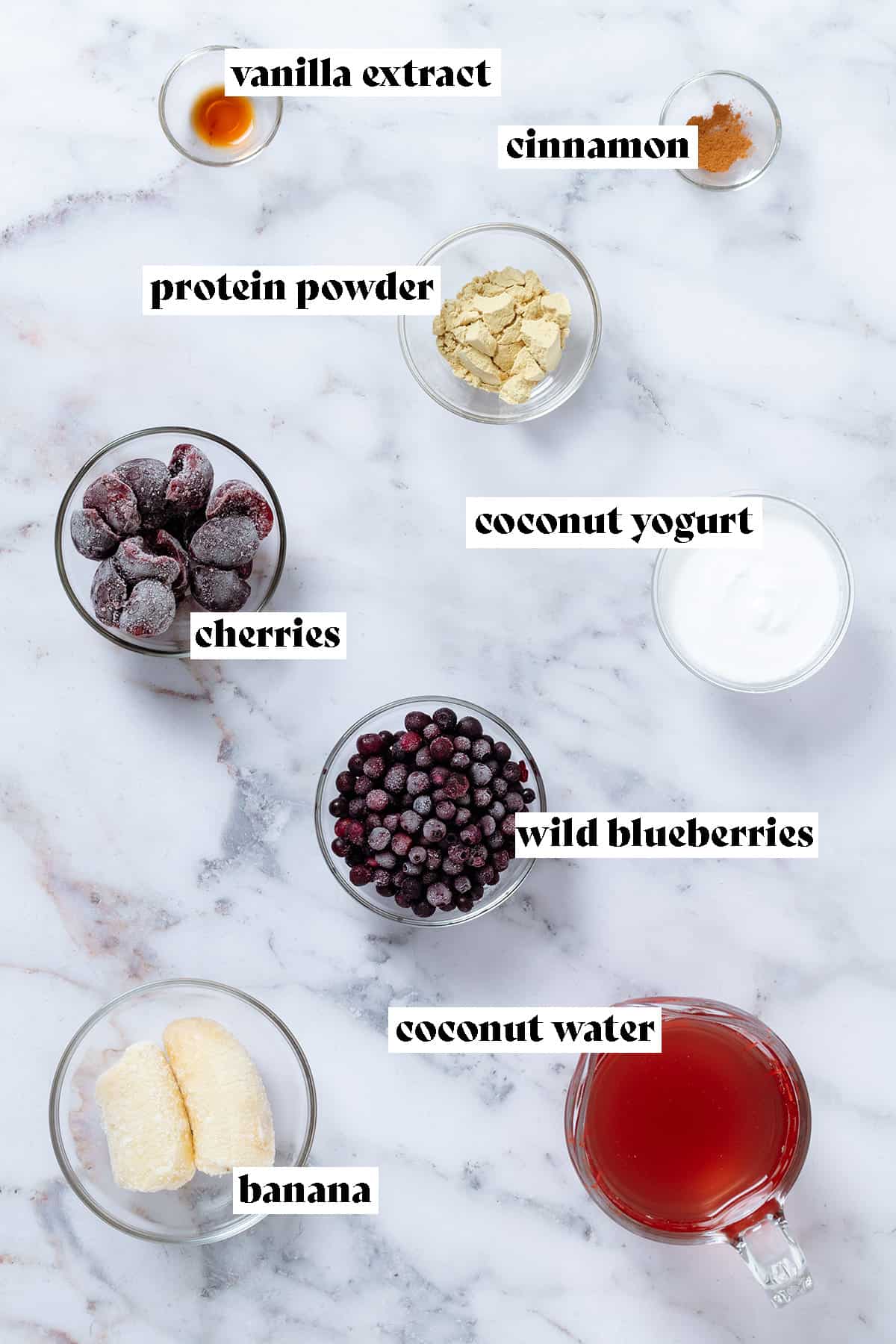 Ingredients in glass bowls like frozen blueberries, cherries, yogurt, banana, protein, and coconut water in a glass measuring cup with text overlay.