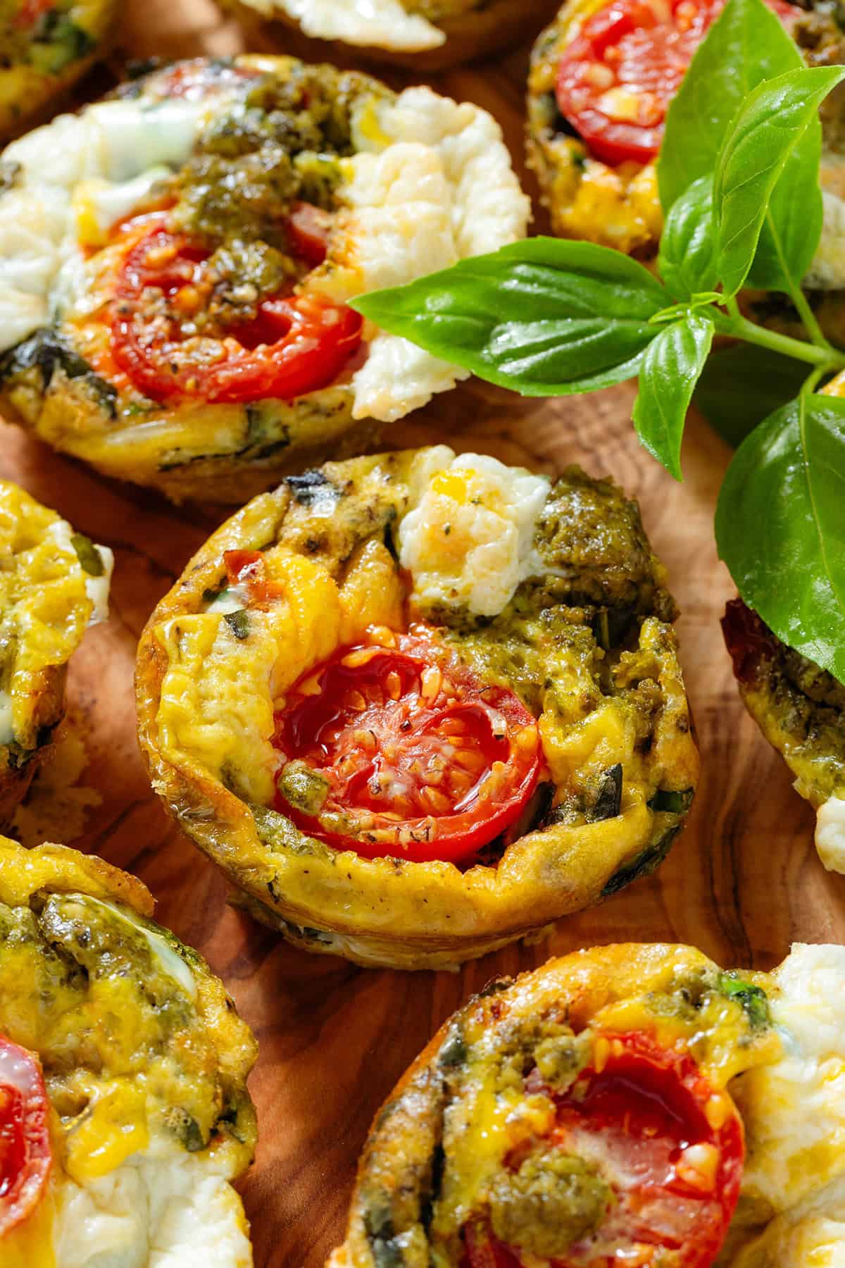 Egg bites with mozzarella, pesto, cherry tomatoes and other ingredients served on a wooden serving board garnished with fresh basil.