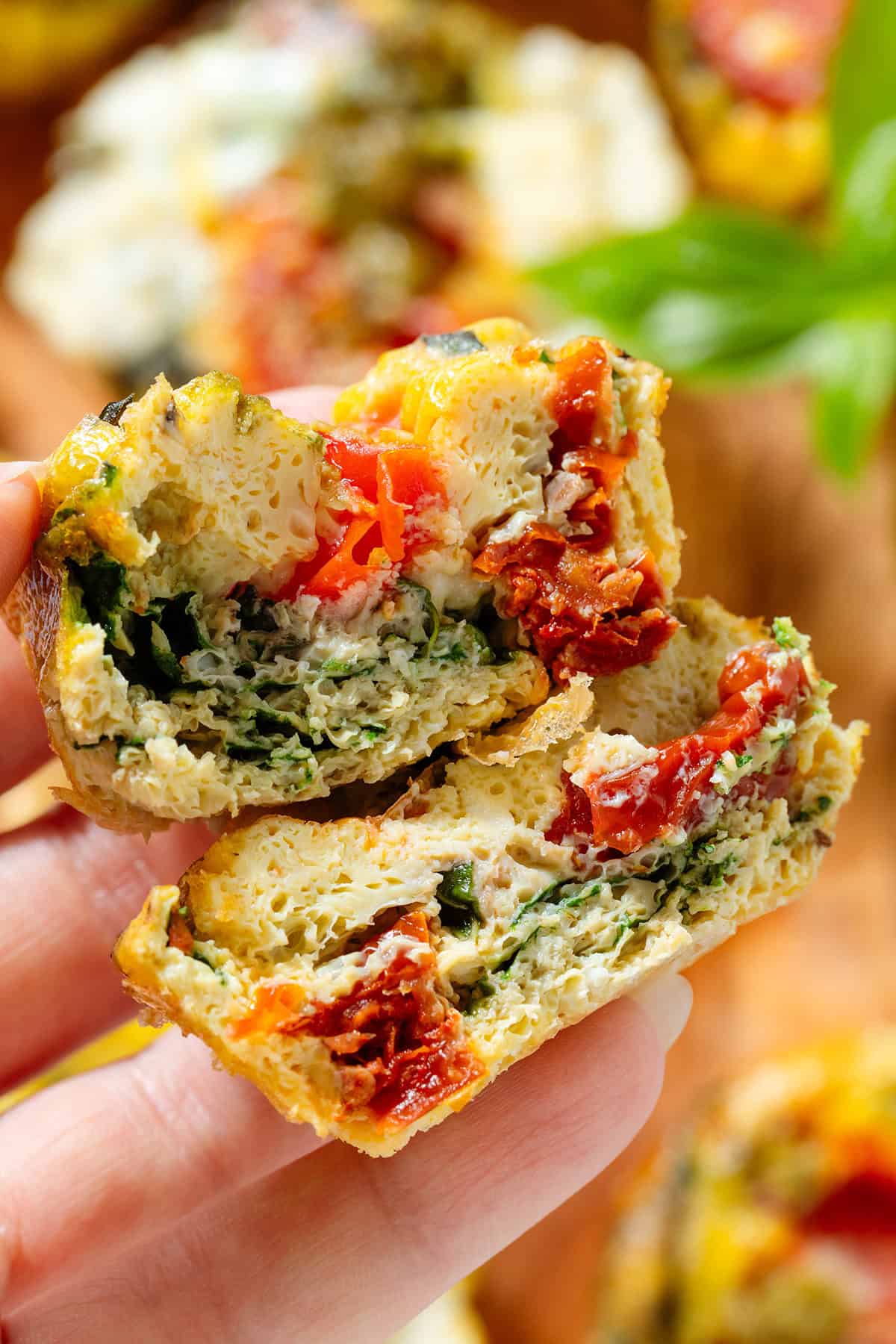 A hand holding an egg muffin sliced in half showing the airy egg, spinach, cherry tomatoes, and other ingredients.