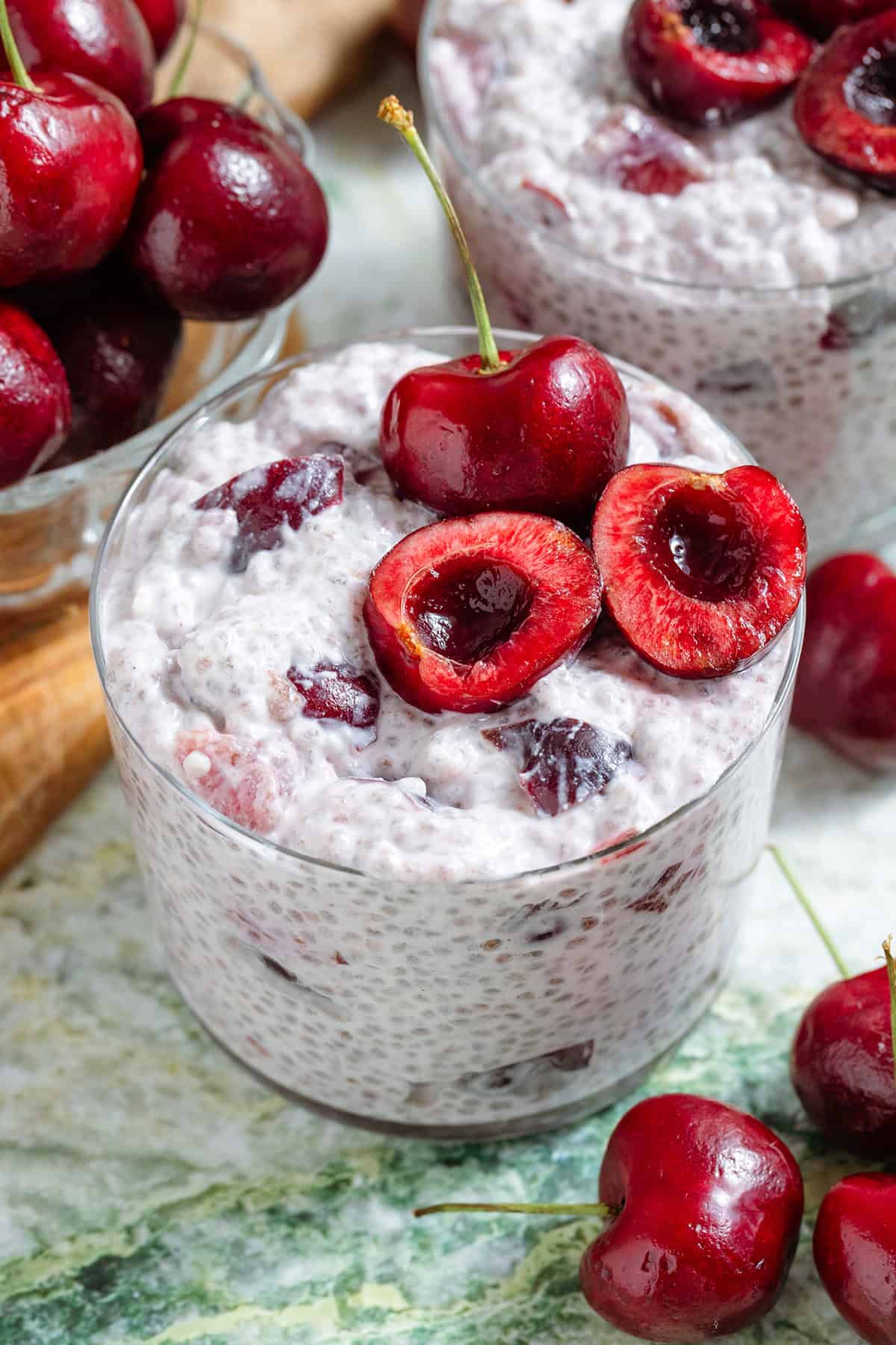 Cherry chia pudding in a short glass garnished with one whole cherry and one cherry sliced in half and pitted and with more cherries around the glass in a small bowl on the left.