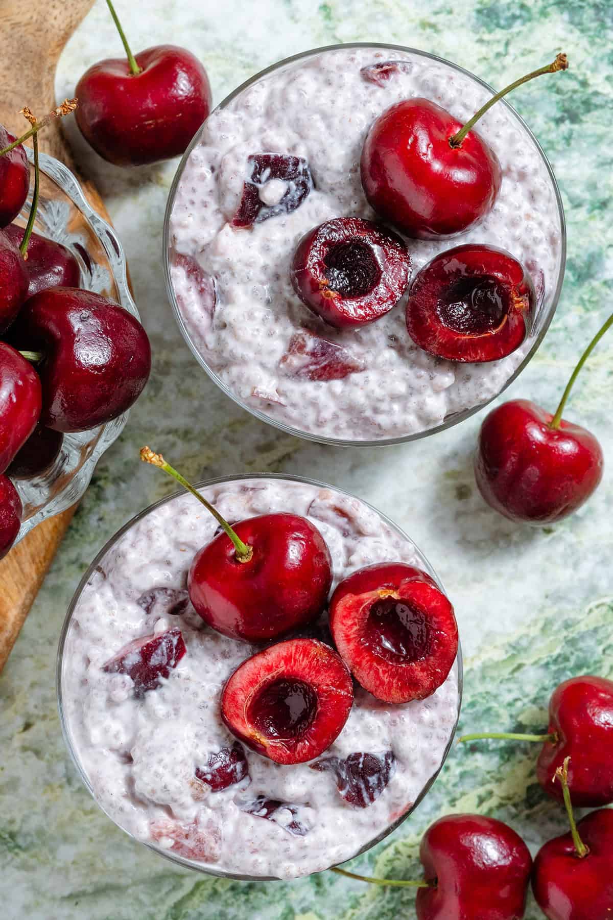 Cherry chia pudding in two short glasses garnished with one whole cherry and one cherry sliced in half and pitted and with more cherries around the glasses.