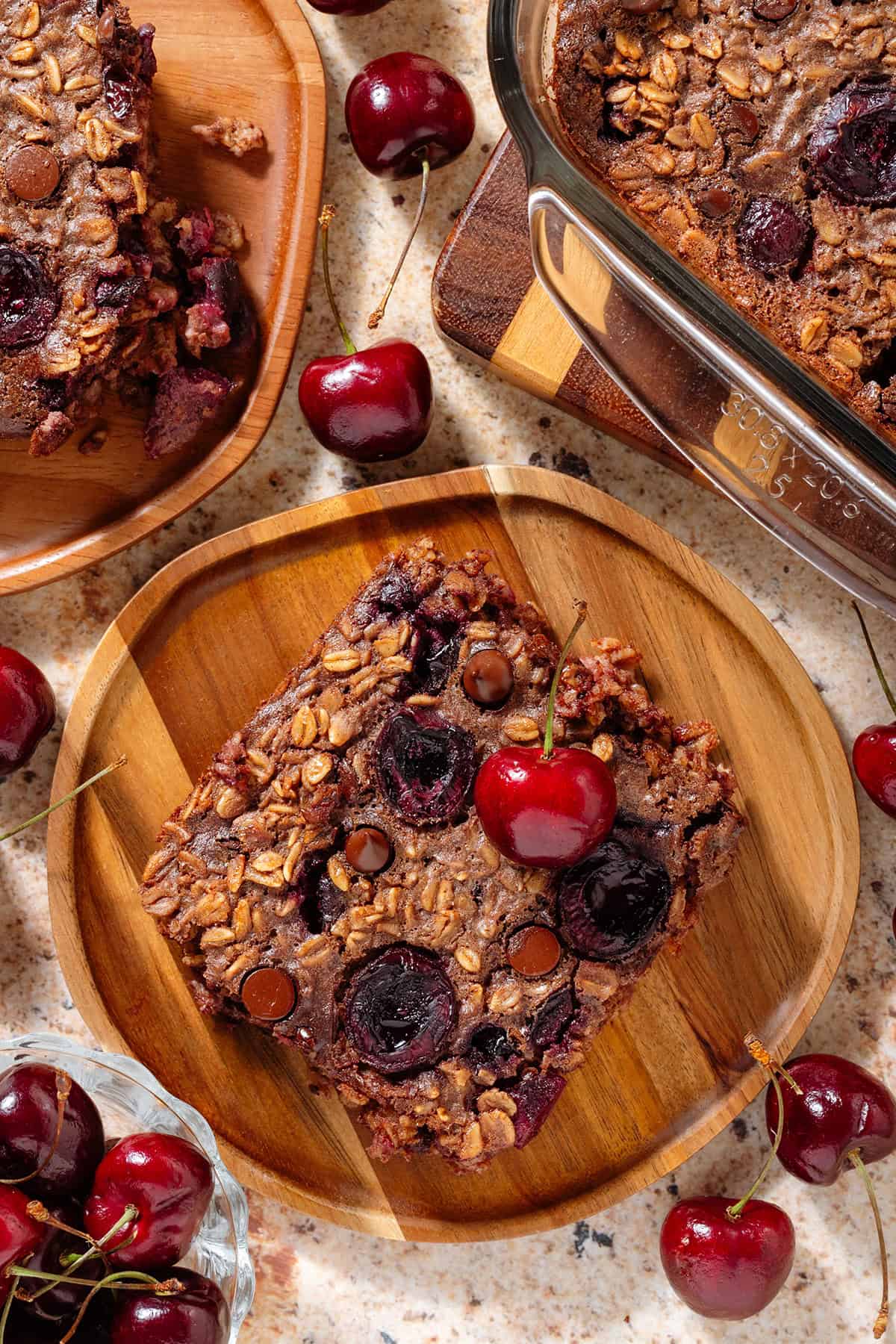 Two slices of a chocolate cherry baked oatmeal on small wooden plates garnished with a fresh cherries.